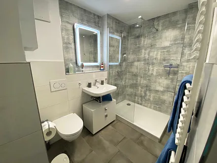 Badezimmer mit offener Glasdusche (niedriger Einstieg), Waschplatz, WC, Kosmetikspiegel, Fön sowie einem Handtuchtrockner und Fußbodenheizung 