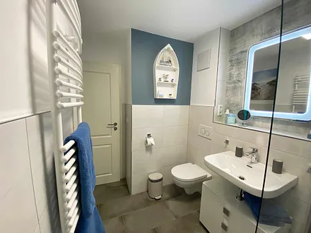 Badezimmer mit offener Glasdusche (niedriger Einstieg), Waschplatz, WC, Kosmetikspiegel, Fön sowie einem Handtuchtrockner und Fußbodenheizung 