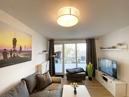 Wohnbereich mit gemütlicher Couch mit Ottomane, Couchtisch, Leselampe, kleines Highboard sowie TV-Board mit Smart-TV und DAB-Radio mit Bluetooth, USB und AUX