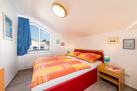 Das Schlafzimmer mit gemütlicher Dachschräge und Doppelbett sowie Gardine und Plissee am Fenster. 