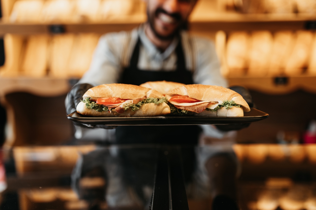 Person in apron holding tray with two fresh sandwiches in a bakery setting.