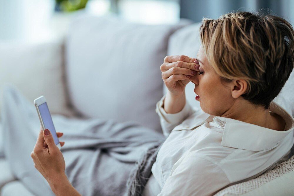 mulher com dor de cabeça segurando a cabeça de dor enquanto relaxa no sofá e usa o celular