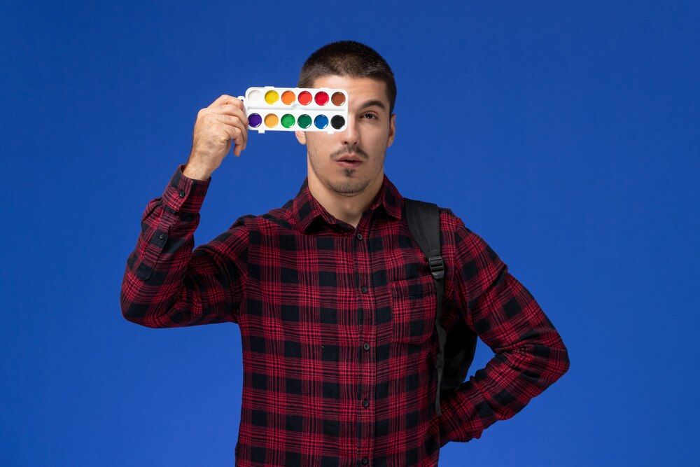 vista frontal de um estudante de camisa xadrez vermelha com mochila segurando tintas para desenho em uma parede azul