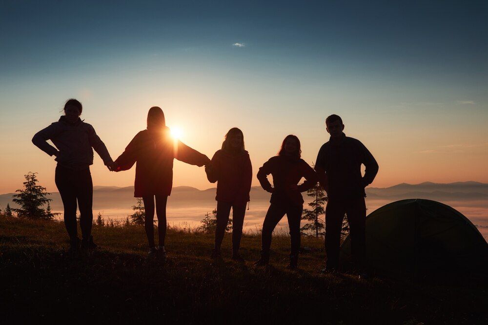 una silueta de un grupo de personas divirtiéndose en la cima de la montaña cerca de la tienda de campaña durante el atardecer.