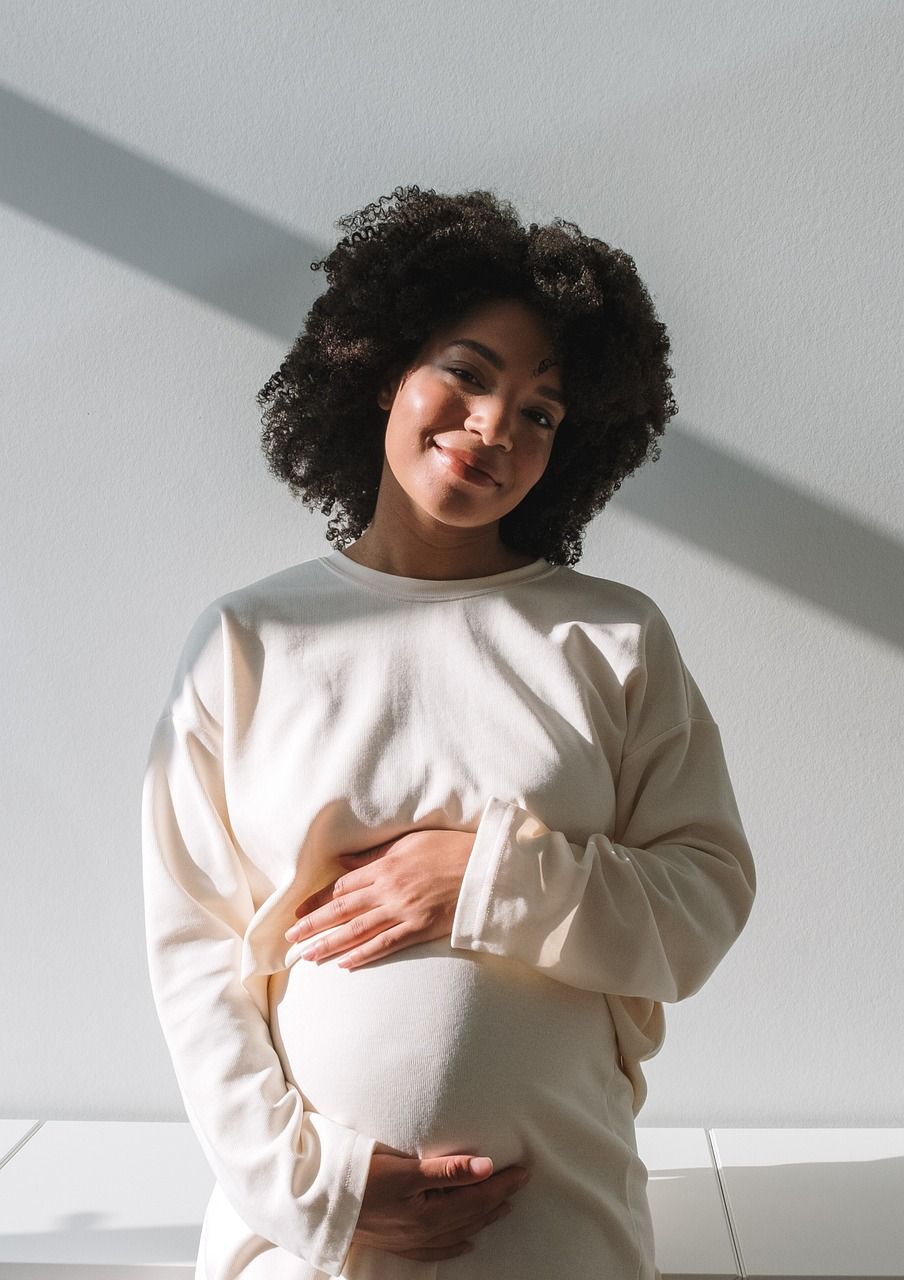 Zu sehen ist eine schwangere Frau mit schwarzen lockigen Haaren in einem weissen Pullover, die Ihren Bauch mit Ihren Händen umrahmt.