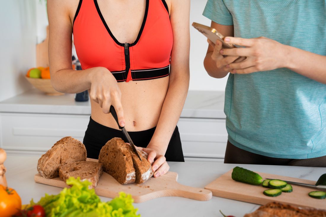 mujer top naranja cortando pan encimera cocina hombre con movil