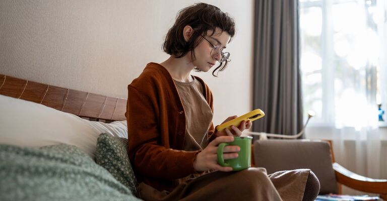 Ragazza sul letto che beve da una tazza e sta al telefono per conto suo