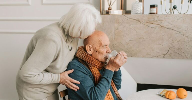 Due anziani, uno seduto e a moglie in piedi che gli bacia la testa