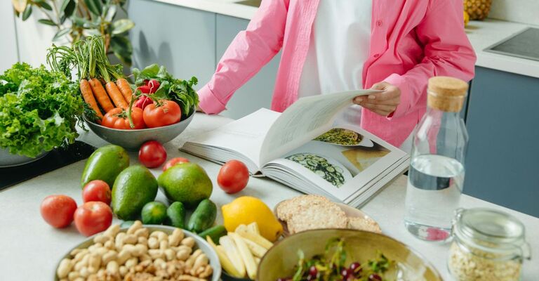 Donna che si accinge a preparare una ricetta a base vegetale. Tavolo con verdure, legumi e lei che sfoglia un ricettario
