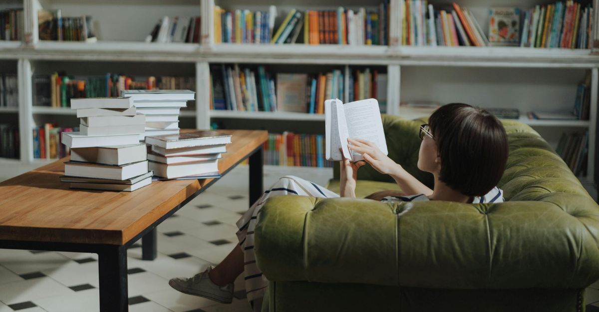 Donna che legge sul divano con davanti una pila di libri sul tavolinetto e una libreria nella stanza