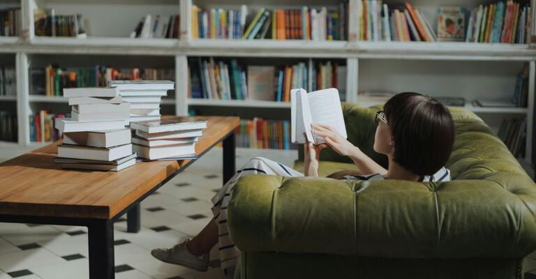 Donna che legge sul divano con davanti una pila di libri sul tavolinetto e una libreria nella stanza