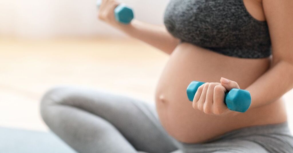 Primo piano di pancia di una donna incinta che si allena a casa con due pesi, uno per mano