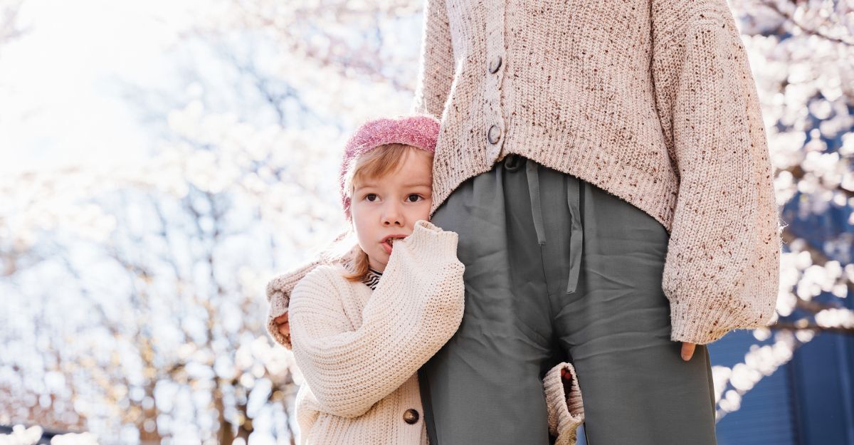 Bambina con il dito in bocca che si attacca alle gambe della mamma