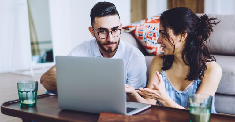 Coppia seduta per terra che osserva il laptop insieme; lei sembra vantarsi con lui di qualcosa