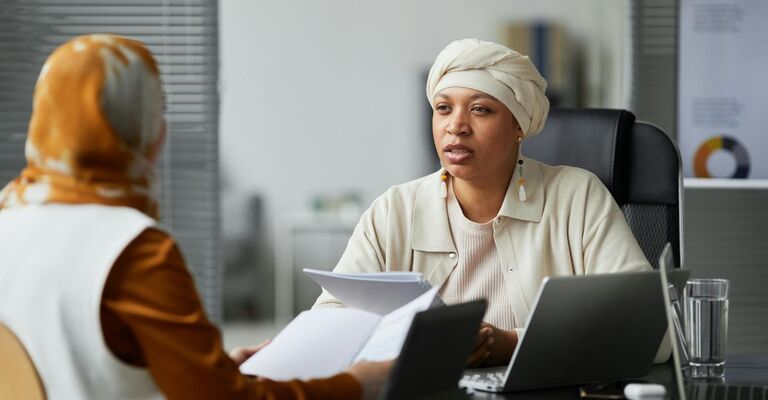 Donna con copricapo che parla con un'impiegata con dei fogli in mano
