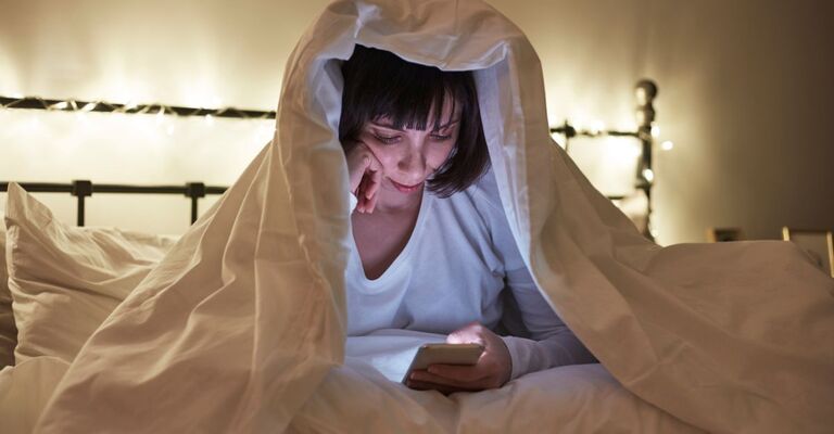 ragazza legge messaggio al cellulare, sotto al piumone