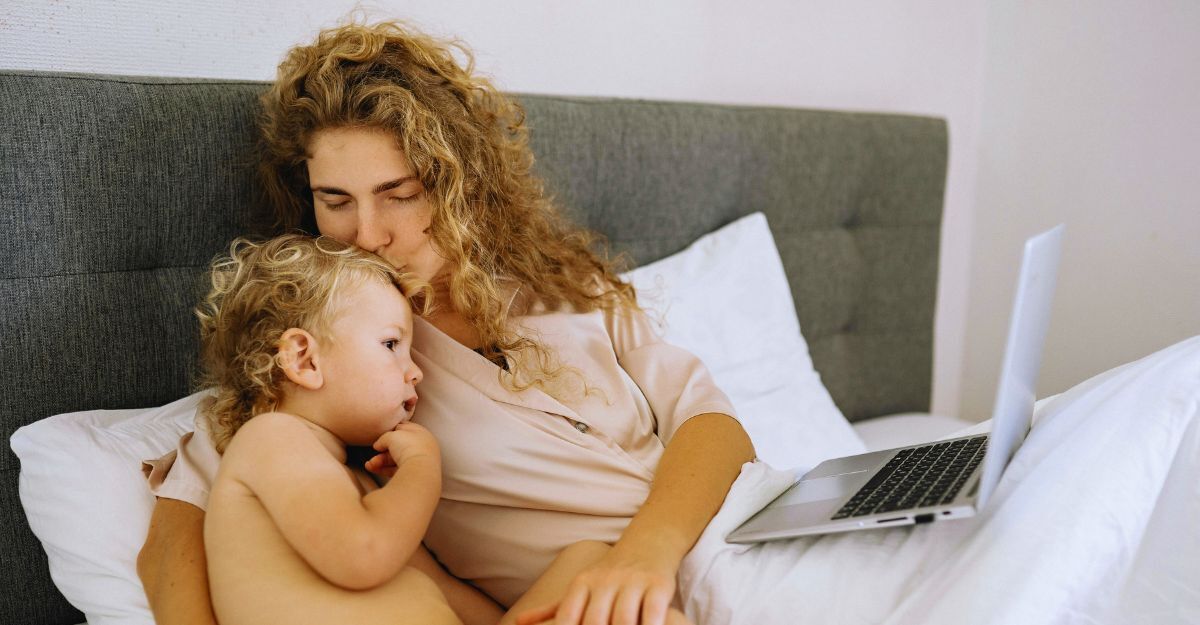 Mamma sdraiata sul letto con il PC sulle ginocchia che dà un bacio sulla testa al figlio neonato