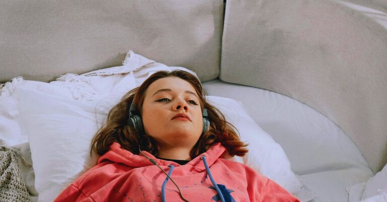 Ragazza in felpa con delle cuffie sulle orecchie che appare riflessiva mentre è sdraiata sul divano