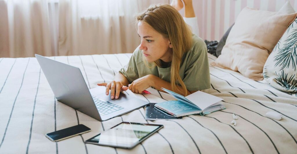 Ragazza sdraiata sul letto che legge al PC, mentre ha sul letto anche tablet, smartphone e un quaderno
