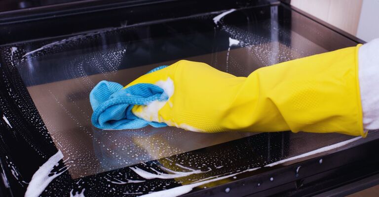 Primo piano di un braccio con guanto giallo e panno in microfibra che spalma uno spray sul vetro del forno per pulirlo