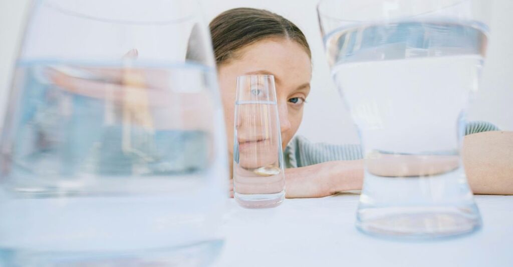 Foto artistica di ragazza seduta su un tavolo con le braccia e bicchieri di varie dimensioni davanti al suo volto che cambiano il suo viso