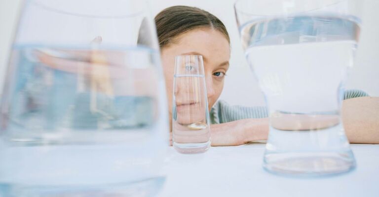 Foto artistica di ragazza seduta su un tavolo con le braccia e bicchieri di varie dimensioni davanti al suo volto che cambiano il suo viso