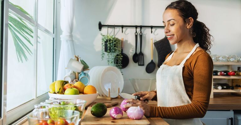 Ragazza che taglia delle verdure davanti alla finestra in cucina, mentre indossa un grembiule