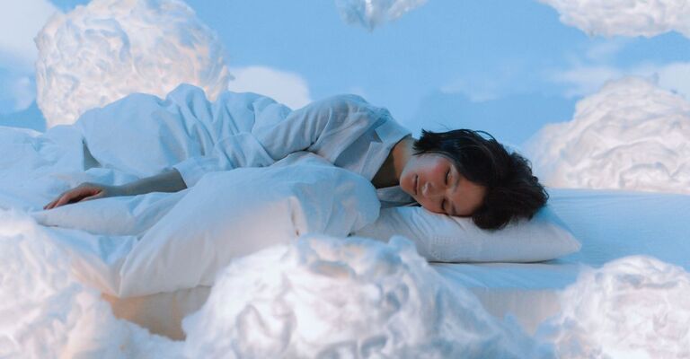 Ragazza sdraiata per terra con la faccia su un cuscino e circondata da nuvole all'interno di un set fotografico