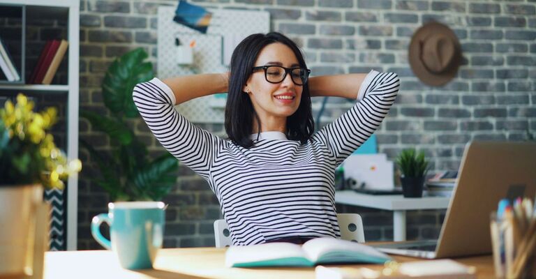 Donna sorride davanti al PC sulla sua scrivania con le braccia dietro la testa, rilassata