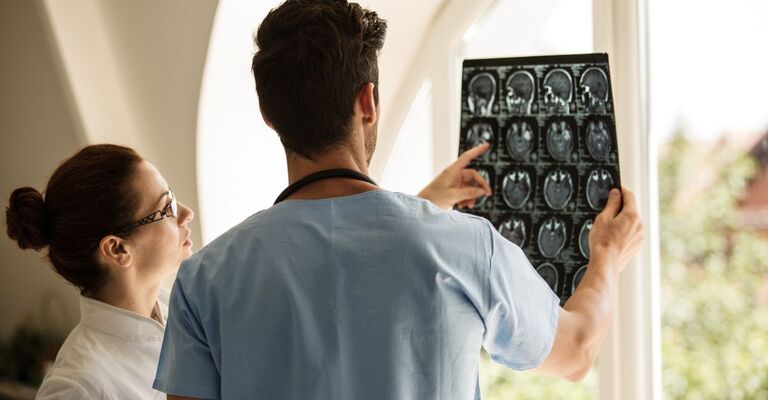 Due medici osservano e analizzano insieme una serie di immagini di risonanza magnetica cerebrale, esaminate alla luce di una finestra.