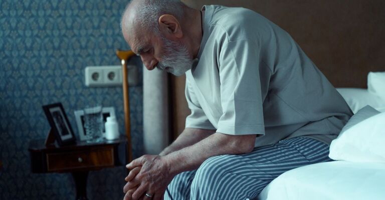 Uomo anziano seduto sul bordo del letto con lo sguardo abbassato, espressione triste o affaticata, in un ambiente domestico.