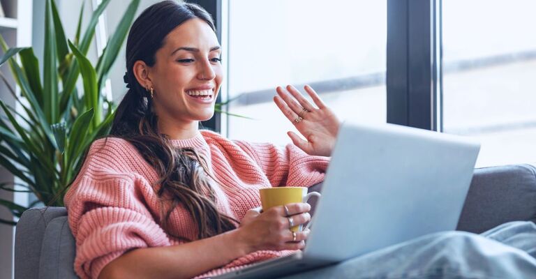 Donna che fa una videochiamata con un'amica sul divano mentre tiene in mano una tazza e sorride