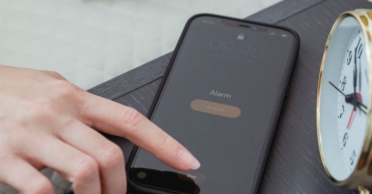 Mano che preme il tasto "Snooze" su uno smartphone per rimandare la sveglia, accanto a una sveglia analogica su un comodino.
