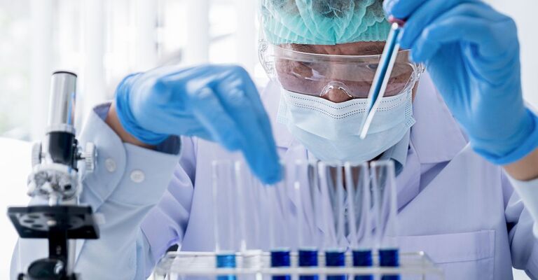 Ricercatore in laboratorio con mascherina, occhiali protettivi e guanti, mentre analizza campioni con una pipetta davanti a una fila di provette.