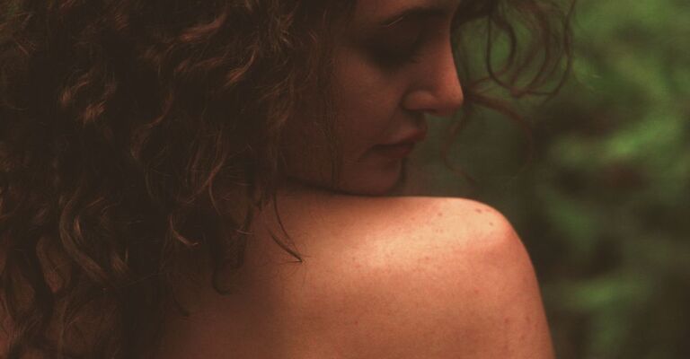 Un primo piano mostra una persona con capelli ricci castani, vista di spalle, che guarda in basso e leggermente a destra. La spalla nuda e la parte superiore della schiena sono visibili, con alcune lentiggini o imperfezioni sulla pelle.