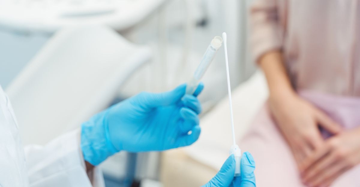 Operatore sanitario con guanti blu tiene un tampone ginecologico sterile, mentre una paziente è seduta sullo sfondo.