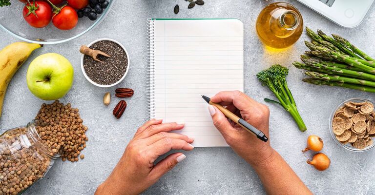 Alimenti di diverso tipo sparsi su un tavolo dove una donna sta scrivendo appunti