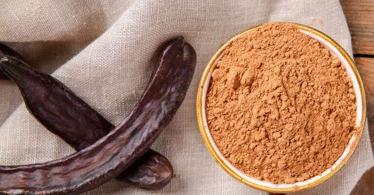 Baccelli secchi di carruba accanto a una ciotola di polvere di carruba su un tessuto di lino.