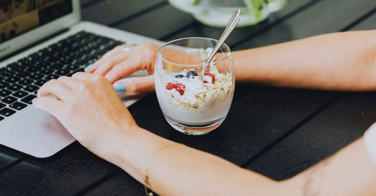 Ragazza al PC che digita con davanti un bicchiere con yogurt, frutta e granola