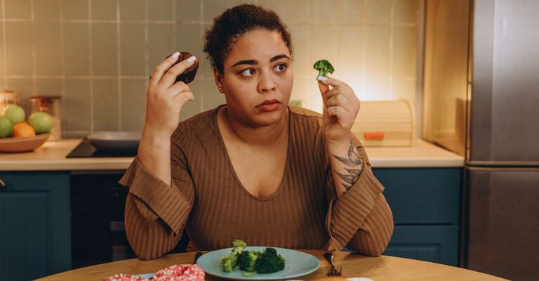 Donna dubbiosa seduta al tavolo con davanti un piatto di broccoli e in mano un broccolo e un muffin