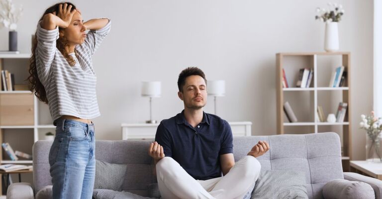 Donna in piedi con le mani sulla testa e uomo seduto sul divano che medita