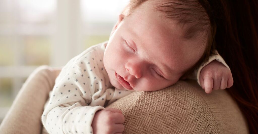 Neonato che dorme serenamente sulla spalla di un adulto, in un ambiente domestico luminoso.