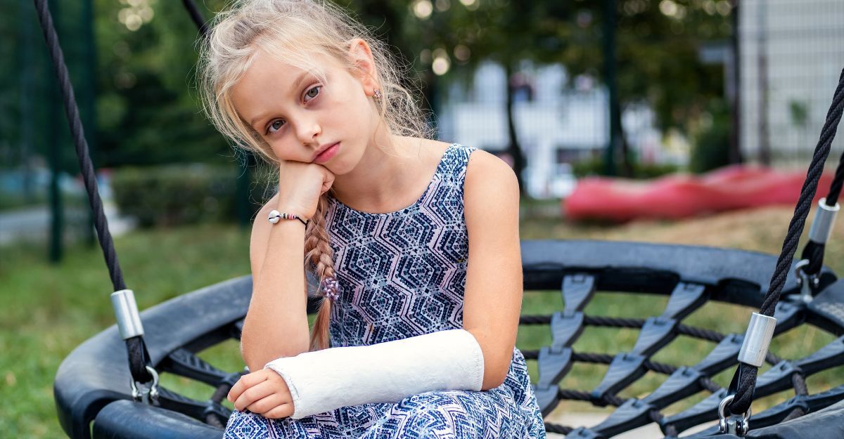 Bambina con braccio ingessato seduta su un'altalena, espressione triste e annoiata dopo una frattura.