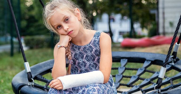 Bambina con braccio ingessato seduta su un'altalena, espressione triste e annoiata dopo una frattura.