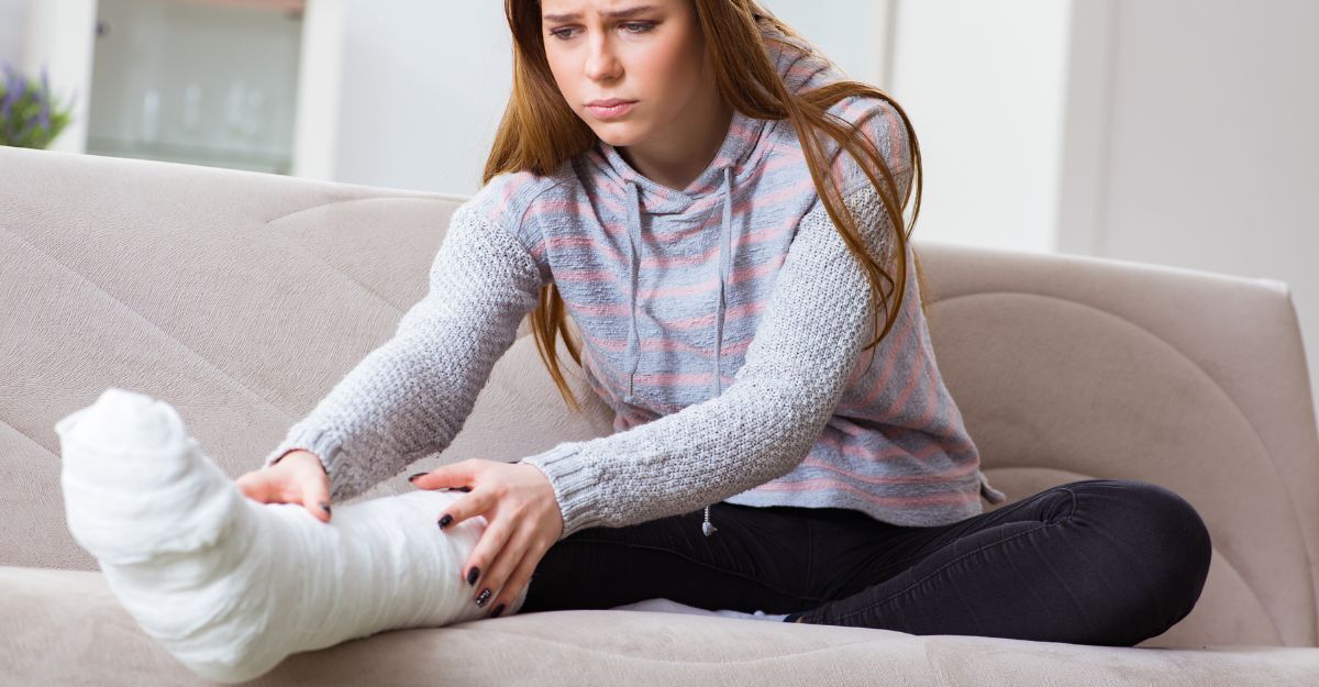 Donna seduta sul divano con gamba ingessata che controlla la frattura, espressione di dolore e preoccupazione.