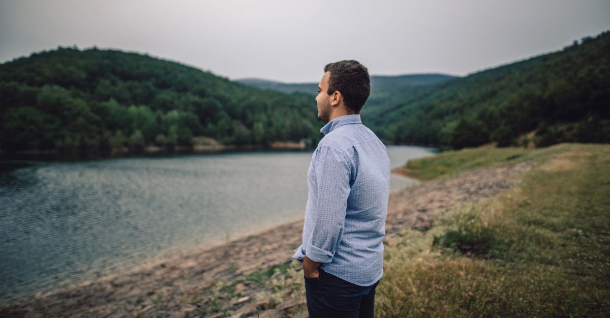 Un uomo guarda un lago con colline boscose sullo sfondo.