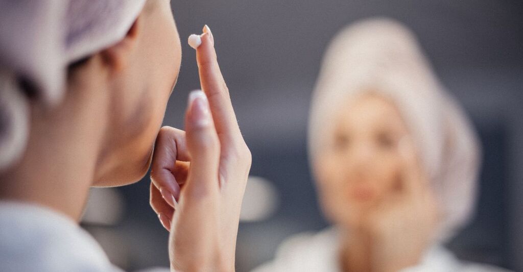 Donna con asciugamano in testa applica crema viso davanti allo specchio