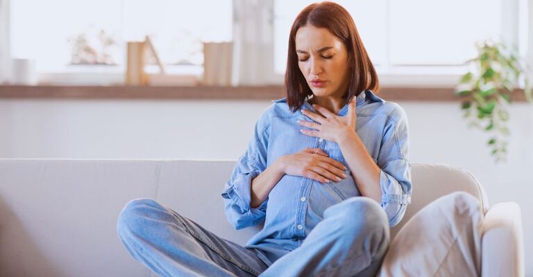 Giovane donna incinta sul divano in jeans e camicia che tiene le mani sul petto mentre è dolorante