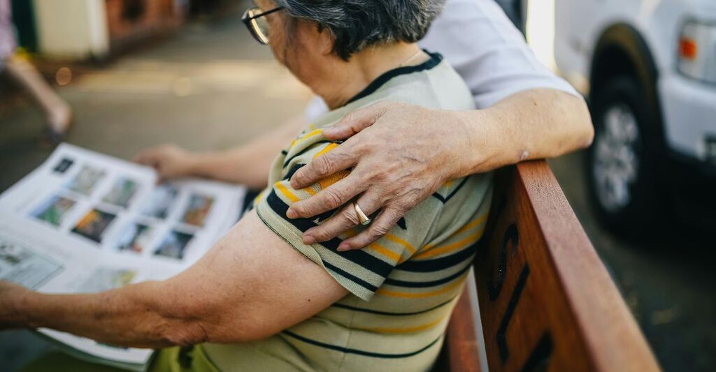 Anziana seduta su una panchina con accanto una persona che la abbraccia affettuosamente mentre legge un giornale