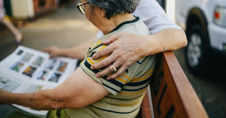 Anziana seduta su una panchina con accanto una persona che la abbraccia affettuosamente mentre legge un giornale